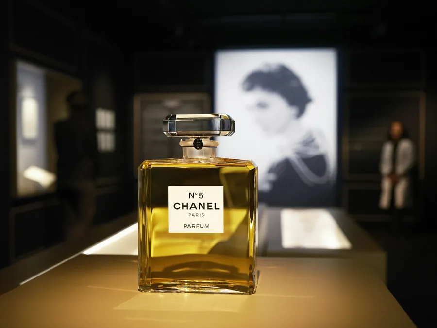 Frasco de CHANEL N°5 iluminado sobre un pedestal, con un retrato en blanco y negro de Gabrielle Chanel detrás