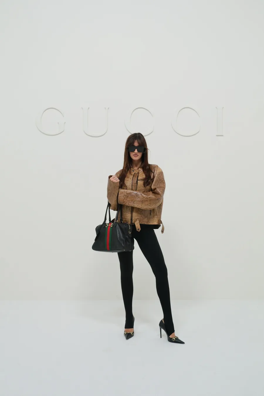 Gucci The Store Show: mujer con chaqueta camel y pantalón negro con un bolso negro de Gucci